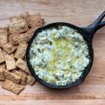 cast iron skillet with spinach artichoke dip on a wooden surface with crackers to the left