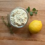 glass bowl with homemade ricotta cheese in it surrounded by a lemon and some herbs