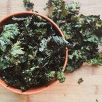 crispy kale chips in an orange bowl with kale chips spilling over onto a wooden surface
